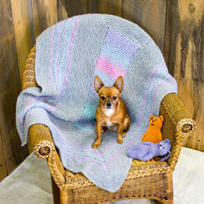 A small dog sits on a wicker chair covered with the Halcyon Yarn's Dream Baby, Baby Blanket download, featuring pastel stripes in a garter stitch design. Next to the dog on the chair are two knitted stuffed animals, one resembling a bear and the other a cat. The background, with its wooden paneling, adds a rustic charm to this cozy scene.