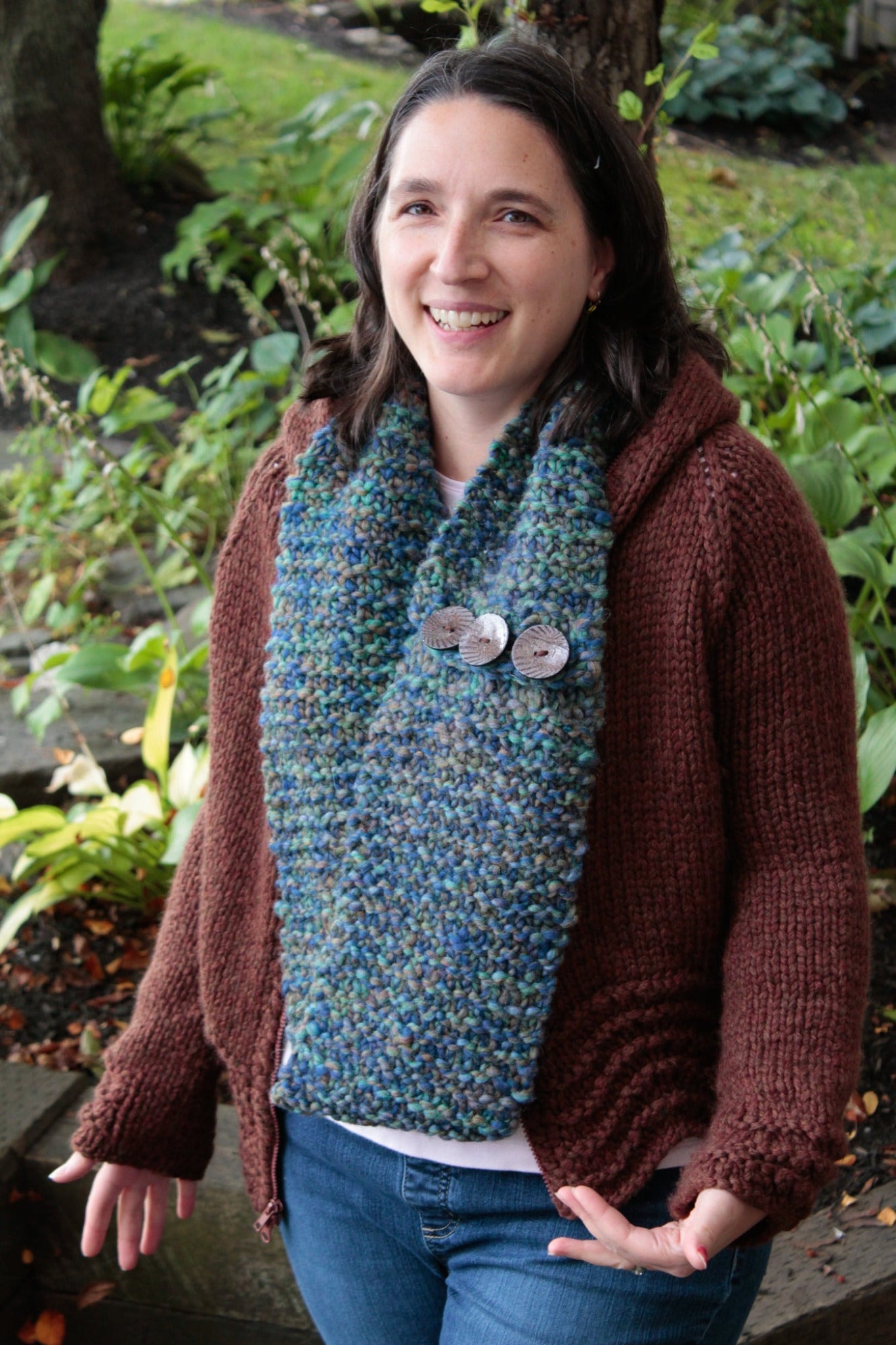 A woman is standing outside, smiling. She is wearing a brown knitted sweater and The Big Easy Cowl by Halcyon Yarn, which has large buttons and a striking blue-green color. The background features lush greenery and trees.