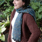 A person with shoulder-length dark hair is standing outdoors, wearing a brown knitted sweater and a blue-green textured Halcyon Yarn's The Big Easy Cowl made of super bulky yarn. They are looking to the left and smiling. The background features lush green plants.