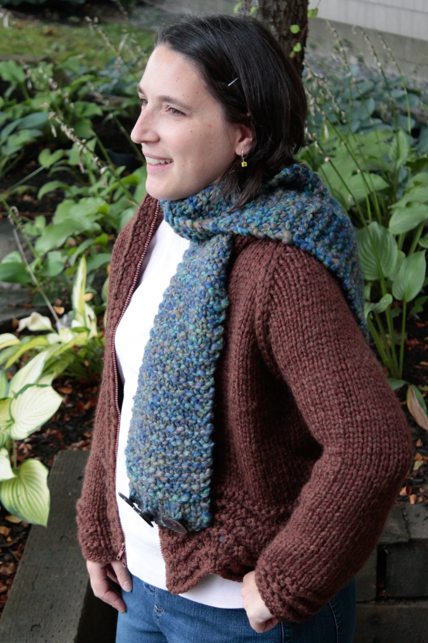 A person with shoulder-length dark hair is standing outdoors, wearing a brown knitted sweater and a blue-green textured Halcyon Yarn's The Big Easy Cowl made of super bulky yarn. They are looking to the left and smiling. The background features lush green plants.