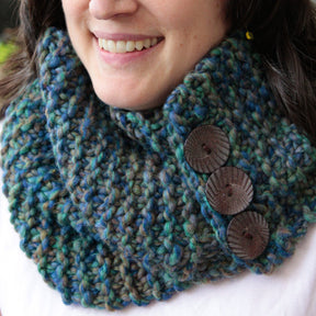 A person wearing The Big Easy Cowl by Halcyon Yarn, a hand-knit, multicolored chunky cowl with three large brown buttons on the right side. The cowl features shades of blue, green, and brown made from super bulky yarn. The person is smiling and wearing a light-colored shirt.