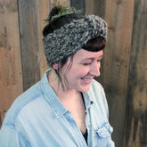 A person with short dark hair and bangs smiles while looking down, wearing the "Head On - Earwarmer" from Halcyon Yarn. This chunky gray knit headband is crafted from super bulky yarn and adorned with a small feather accessory. The individual pairs it with a light denim button-up shirt, set against a backdrop of wooden panels.