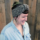 A person with short, dark hair, wearing the "Head On - Earwarmer" from Halcyon Yarn—a chunky headband knitted with super bulky yarn and adorned with a tucked-in feather—smiles while looking downward. They are dressed in a light blue button-up shirt and stand against a backdrop of wooden planks.