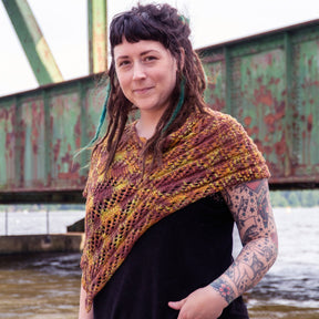 A person with long, dreadlocked hair stands outside near a rusty bridge, adorned in a black shirt and a vibrant Dunes Poncho by Halcyon Yarn. The poncho is crafted from hand-dyed organic cotton and showcases intricate patterns ideal for advanced beginner knitters. Visible tattoos decorate their arms as they offer a slight smile to the camera, with water visible in the background.