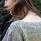 A person with shoulder-length hair looks down, showcasing the upper back and shoulder details of a Halcyon Yarn Hootenanny Sweater. The sweater, crafted from heavy worsted yarn, features a textured pattern in shades of green and gray. The green background is out of focus.