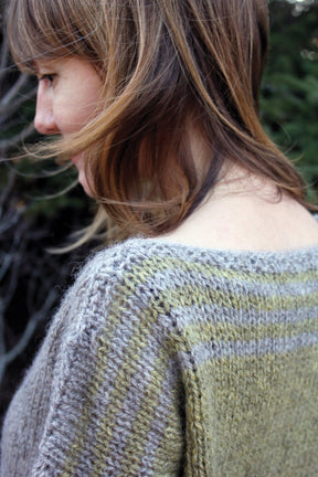 A person with shoulder-length hair looks down, showcasing the upper back and shoulder details of a Halcyon Yarn Hootenanny Sweater. The sweater, crafted from heavy worsted yarn, features a textured pattern in shades of green and gray. The green background is out of focus.