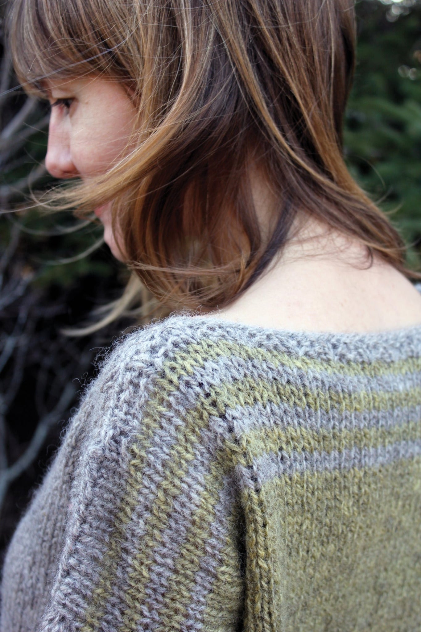 A person with shoulder-length brown hair stands sideways, showcasing the back of their Hootenanny Sweater from Halcyon Yarn. The sweater features green and gray stripes, and the blurred background hints at foliage. The lighting highlights the texture and stitches of the women's pullover.