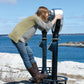 A person wearing a Hootenanny Sweater by Halcyon Yarn, along with jeans and brown boots, is standing on the platform of a coin-operated binocular viewer at a seaside location, looking through the binoculars. The background showcases a body of water and a distant shoreline under a blue sky.