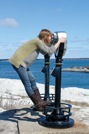 A person wearing the Hootenanny Sweater by Halcyon Yarn in gray and green, paired with jeans and brown boots, stands on tiptoe while peering through a coin-operated binocular viewer. The viewer is positioned in a coastal area where the ocean and rocky shoreline are visible in the background.