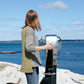 A person with shoulder-length hair, wearing the green and gray Hootenanny Sweater by Halcyon Yarn and jeans, looks through a coin-operated binocular viewer. They stand on a rocky surface overlooking a blue ocean under a partly cloudy sky.