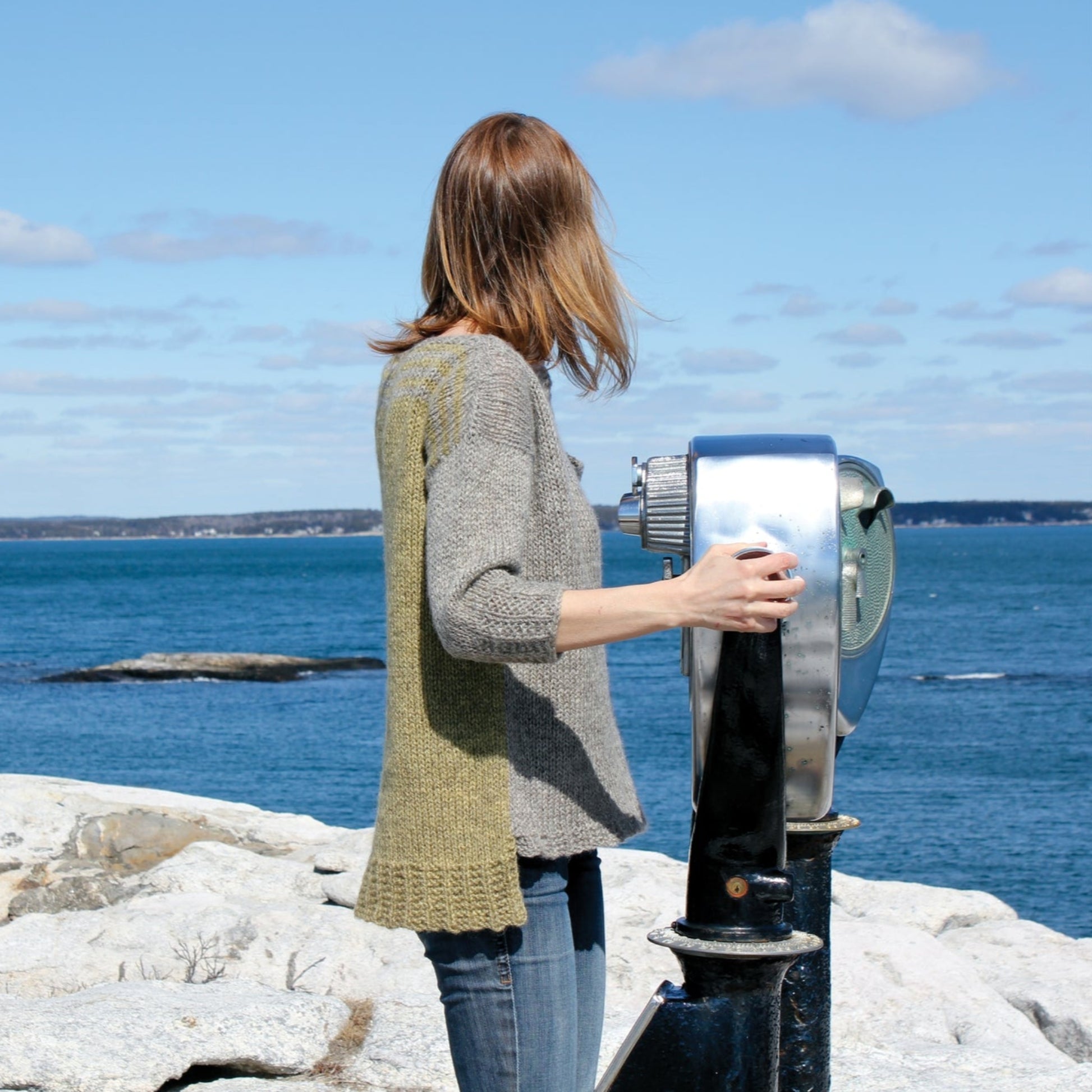 A person with shoulder-length brown hair, wearing the grey and green Hootenanny Sweater by Halcyon Yarn, is looking out at the ocean through a telescope viewer. They stand on rocky terrain with the blue sea and a partly cloudy sky in the background.