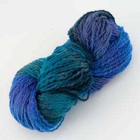 A skein of Great Adirondack Galloway hand-dyed merino yarn from Great Adirondack Yarn Co, Inc. features variegated blue and teal tones. The yarn is twisted, showcasing a gradient that transitions smoothly between different shades, giving a textured appearance. Perfect for creating unique accessories, the background remains plain white.