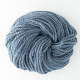 A close-up image of Baby Alpaca Grande by Plymouth yarn in a light denim blue color, tightly wound and coiled into a neat bundle. The ultra-soft Plymouth Yarn Co. yarn appears thick and bulky weight, perfect for knitting or crocheting projects. The background is plain white.