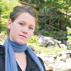 A woman with brown hair tied back, wearing an elegant blue top and the Halcyon Yarn Romantic Silk and Mohair Scarf, stands outdoors in a natural setting with greenery and rocks in the background. She has a calm, neutral expression on her face.