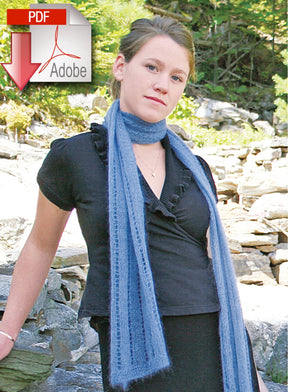 A person standing outdoors on a rocky landscape, wearing a black top and the Romantic Silk and Mohair Scarf in light blue by Halcyon Yarn. A PDF Adobe icon with a red downward arrow is displayed in the top left corner of the image. Green foliage is visible in the background, reminiscent of an evening in Paris.