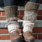 Close-up of a person sitting on outdoor steps, wearing the Down Home Special Leg Warmers by Halcyon Yarn in brown and cream, knitted from Icelandic wool over black pants. The variegated Plötulopi pattern pairs beautifully with their brown leather shoes. The steps are made of concrete with brick risers.