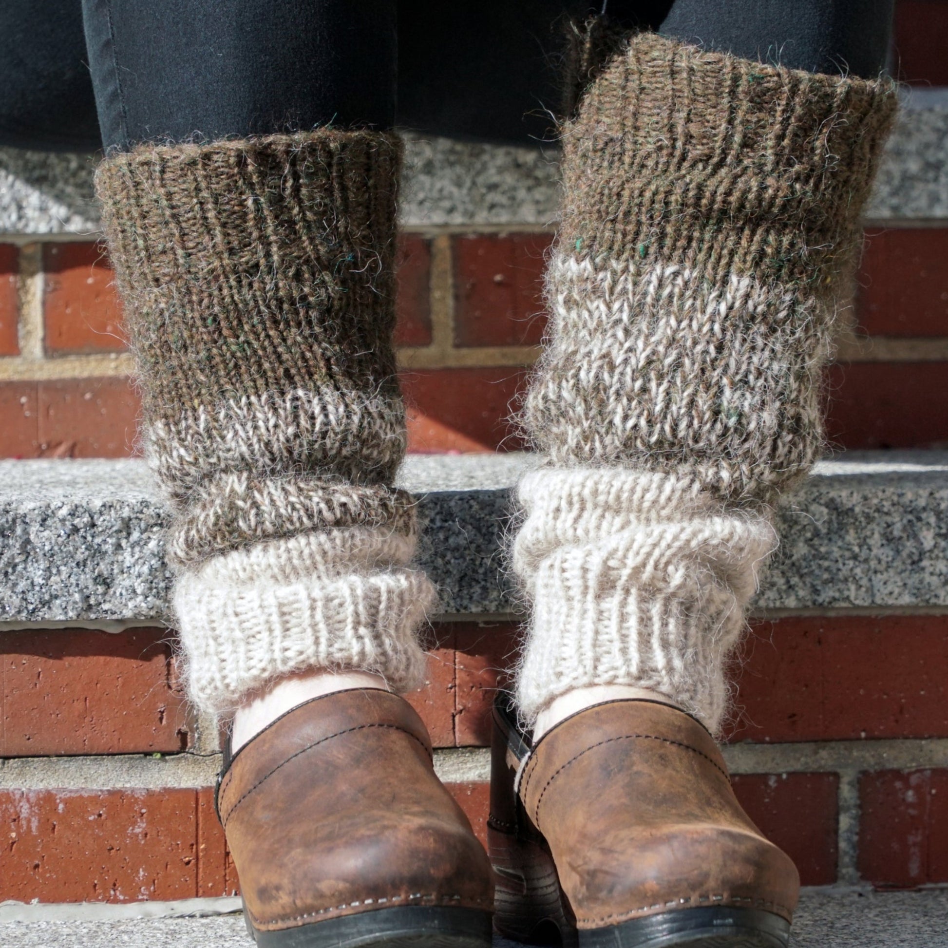 Close-up of a person sitting on outdoor steps, wearing the Down Home Special Leg Warmers by Halcyon Yarn in brown and cream, knitted from Icelandic wool over black pants. The variegated Plötulopi pattern pairs beautifully with their brown leather shoes. The steps are made of concrete with brick risers.