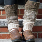 A close-up of a person sporting Down Home Special Leg Warmers from Halcyon Yarn, showcasing their thick, cozy knit made from Icelandic wool. The leg warmers feature a gradient pattern in shades of brown, gray, and white and are layered over black leggings and paired with leather shoes that have wooden soles. The person is standing on concrete steps against a backdrop of a brick wall.