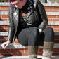 A person with bright purple hair, wearing a black leather jacket adorned with pins, a graphical T-shirt, black jeans, Halcyon Yarn's Down Home Special Leg Warmers, and brown shoes, sits on outdoor brick steps. They look down and rest one hand on the step beside them.