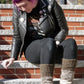 A person with vibrant purple hair sits on outdoor steps. They wear a black leather jacket adorned with pins, black pants, and Halcyon Yarn's Down Home Special Leg Warmers made of cozy Icelandic wool over brown shoes. The person looks down with a slight smile. The background includes a brick wall and greenery.