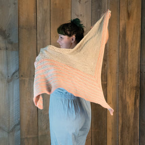 A person wearing a light blue dress stands in front of a wooden wall, holding up the large "Waiting Room Crocheted Shawl" by Halcyon Yarn, with its shades of pink and beige. The person is turned slightly to the side, showcasing the shawl's triangular shape and intricate crochet pattern.