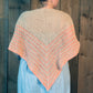 A person is wearing the Waiting Room Crocheted Shawl by Halcyon Yarn, featuring beige and pink stripes over a light blue outfit, standing in front of a rustic wooden wall. The shawl showcases an intricate crochet pattern with a textured chevron design, draping elegantly over the person's back and shoulders.