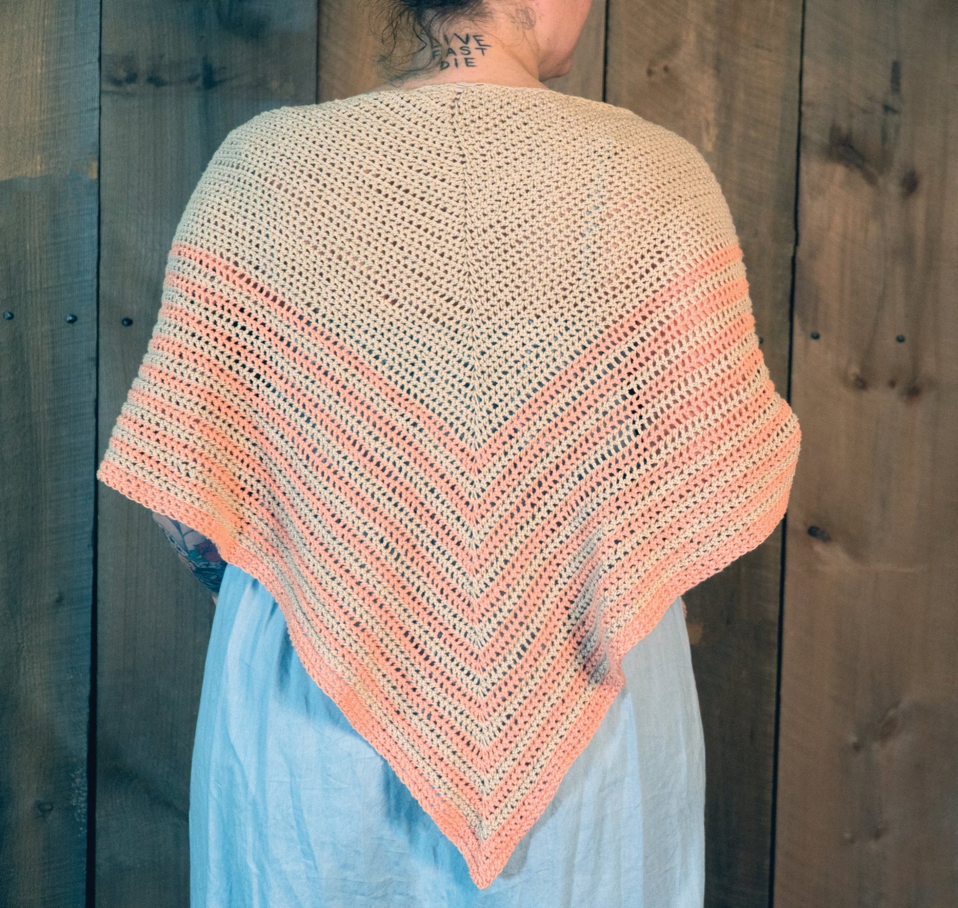 A person is shown from the back wearing a light blue dress and the Waiting Room Crocheted Shawl by Halcyon Yarn, featuring a V-shape design. The shawl has a beige color on top that transitions into peach at the edges. The background features wooden planks.