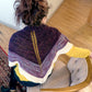 A person with dark hair tied up in a bun is seated on a chair, viewed from the back. They are wearing the Space Oddity Shawl by Halcyon Yarn, which features garter stitch in purple, gray, and white stripes over a yellow top and black pants. The setting appears to be an indoor space with wooden floors and a patterned rug.