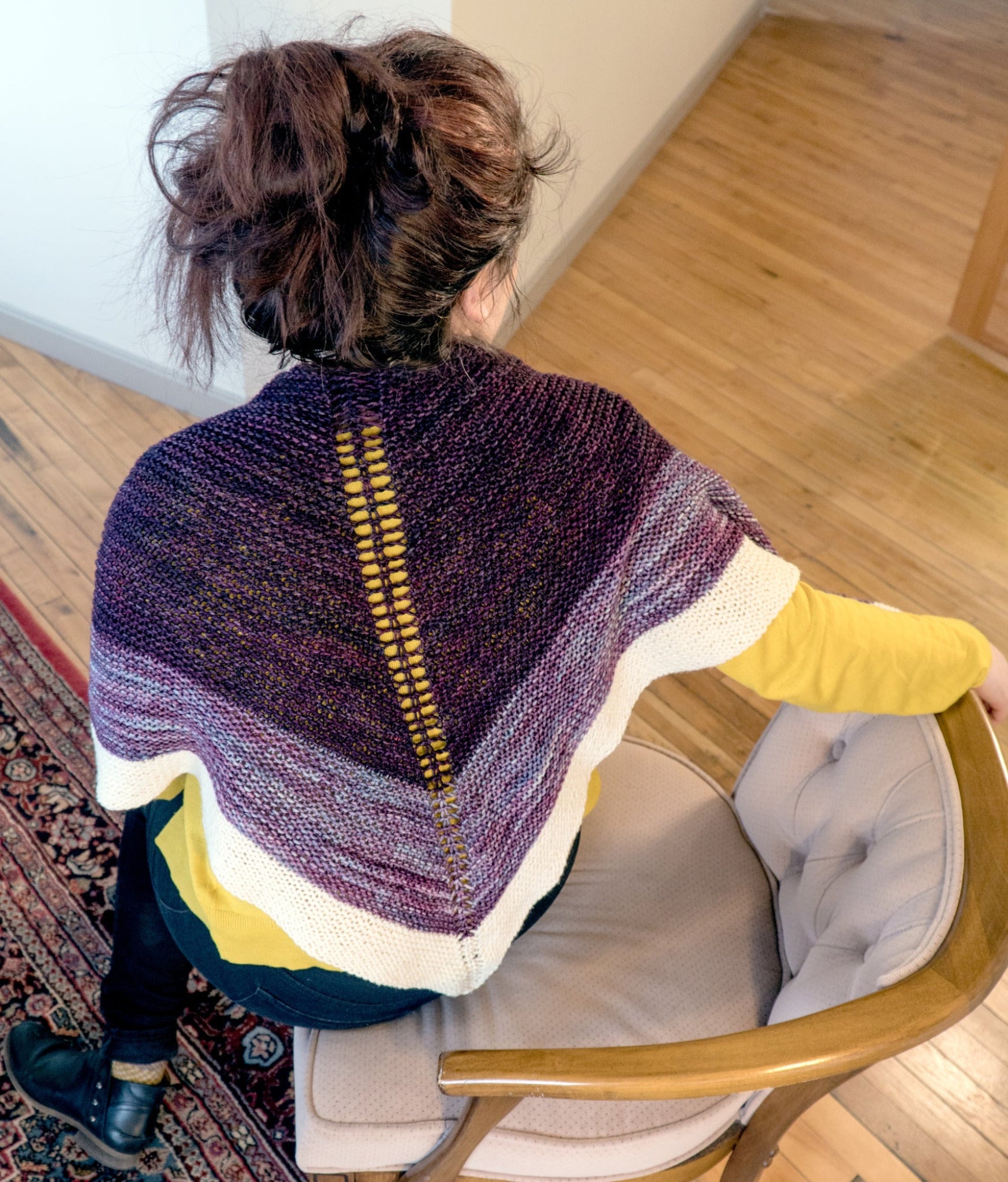 A person with dark hair tied up in a bun is seated on a chair, viewed from the back. They are wearing the Space Oddity Shawl by Halcyon Yarn, which features garter stitch in purple, gray, and white stripes over a yellow top and black pants. The setting appears to be an indoor space with wooden floors and a patterned rug.