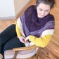 A woman with dark hair sits in a wooden chair on a light-toned hardwood floor. She is wearing a mustard yellow sweater, dark pants, and the Space Oddity Shawl by Halcyon Yarn, crafted from worsted weight yarn in shades of purple, white, and gray. The garter stitch of the shawl adds texture as she looks to her right, appearing thoughtful.