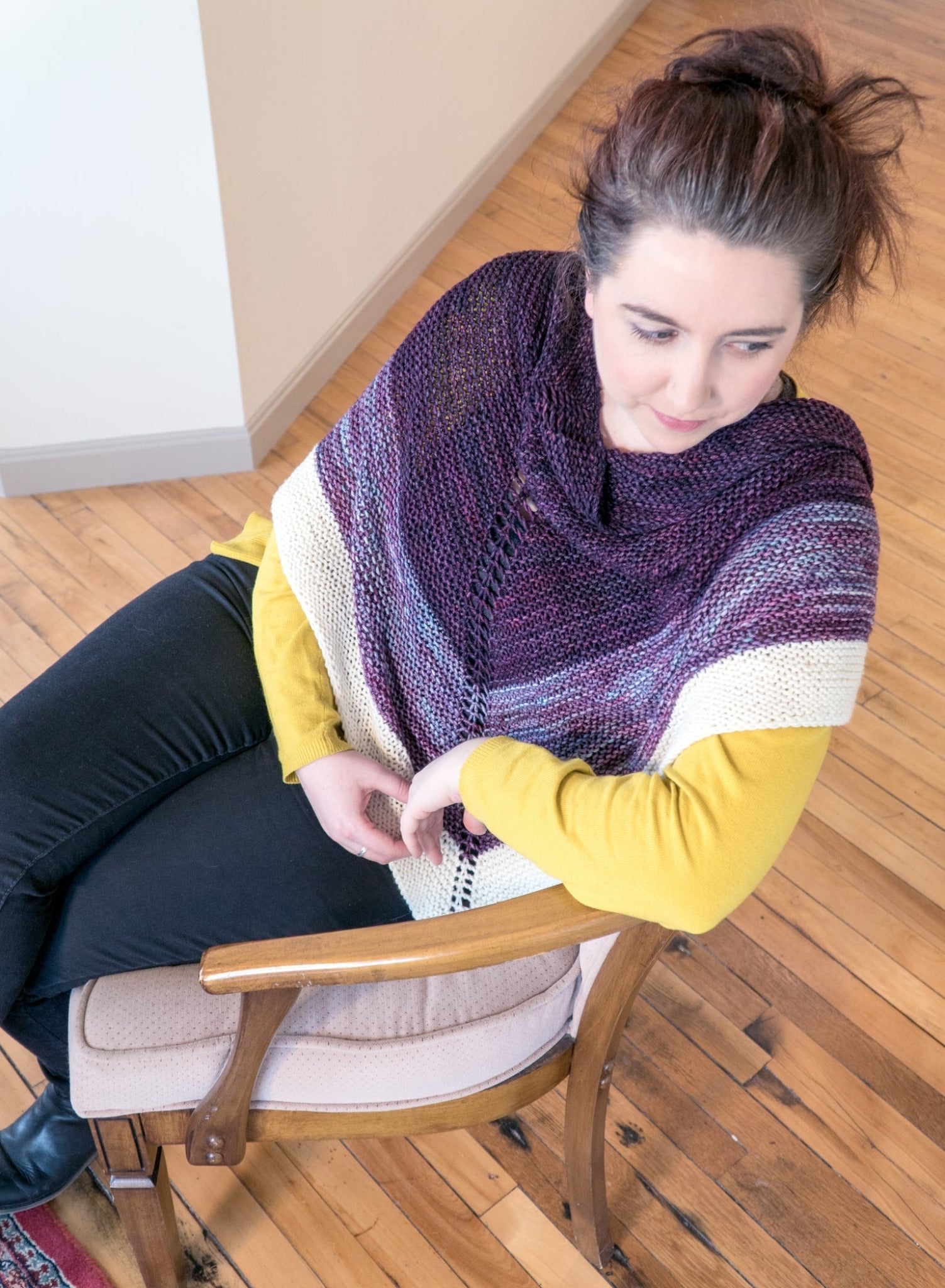 A woman with dark hair sits in a wooden chair on a light-toned hardwood floor. She is wearing a mustard yellow sweater, dark pants, and the Space Oddity Shawl by Halcyon Yarn, crafted from worsted weight yarn in shades of purple, white, and gray. The garter stitch of the shawl adds texture as she looks to her right, appearing thoughtful.