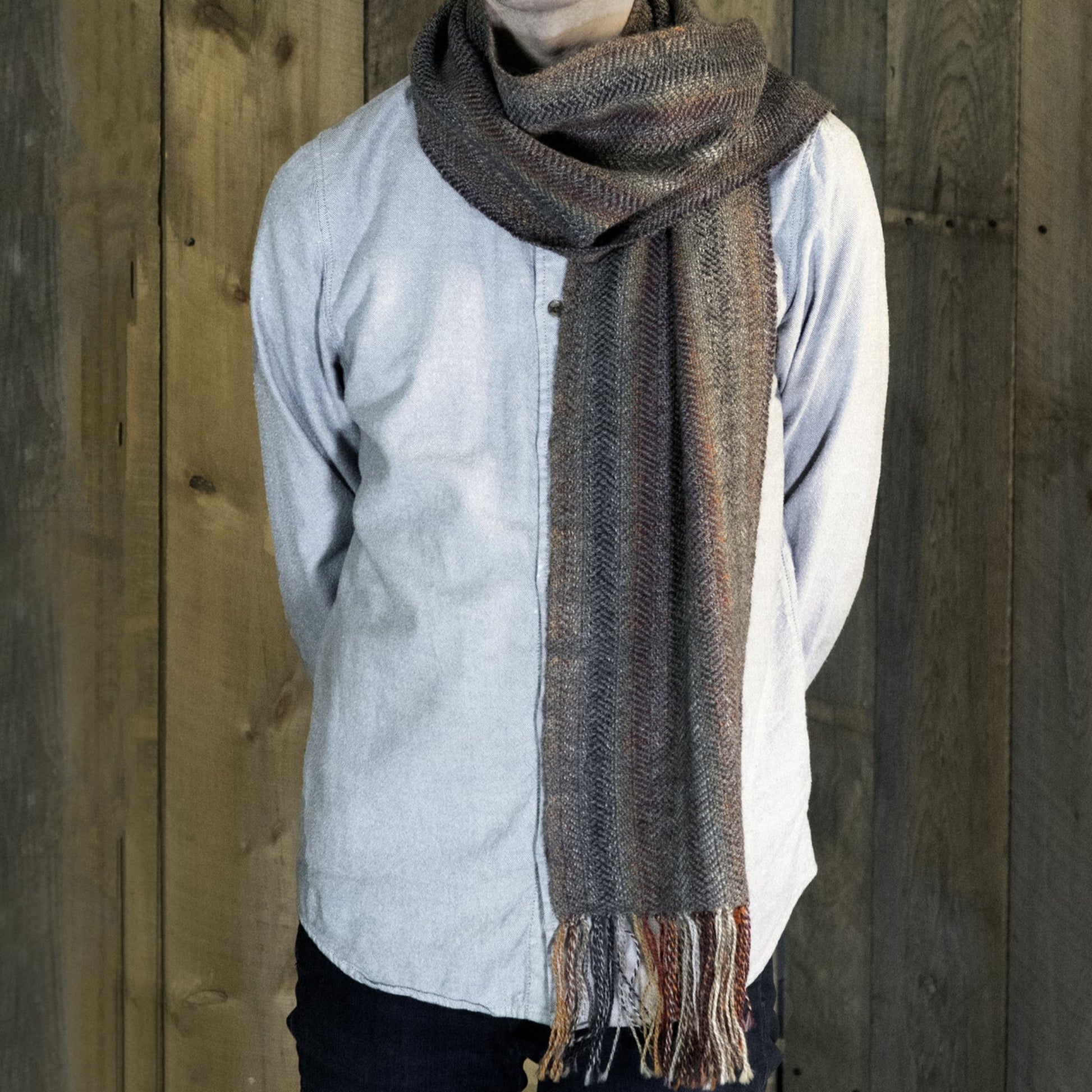 Person wearing a light-colored button-up shirt and a textured gray "Oktoberfest - Woven Scarf" by Halcyon Yarn stands in front of a wooden wall, with their arms behind their back. Only their torso and part of their neck are visible. The scarf has tassels hanging from the ends.