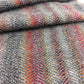 A close-up of the Oktoberfest - Woven Scarf by Halcyon Yarn shows a folded piece of fabric in shades of gray, featuring subtle, multicolored stripes reminiscent of self-striping sock yarn running vertically. The texture appears soft and intricate, showcasing the detailed weaving pattern.