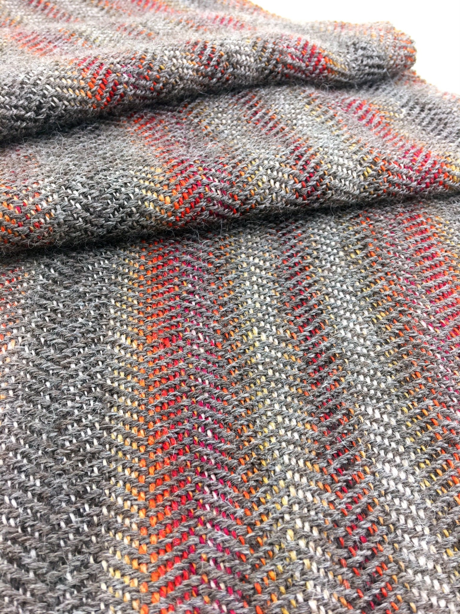 A close-up of the Oktoberfest - Woven Scarf by Halcyon Yarn shows a folded piece of fabric in shades of gray, featuring subtle, multicolored stripes reminiscent of self-striping sock yarn running vertically. The texture appears soft and intricate, showcasing the detailed weaving pattern.