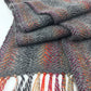 A close-up image of the Halcyon Yarn Oktoberfest - Woven Scarf, crafted from gray Herriot Fine alpaca yarn with delicate accents of orange, red, and purple. The scarf is adorned with a fringe composed of colorful strands at the ends, displayed against a white background.