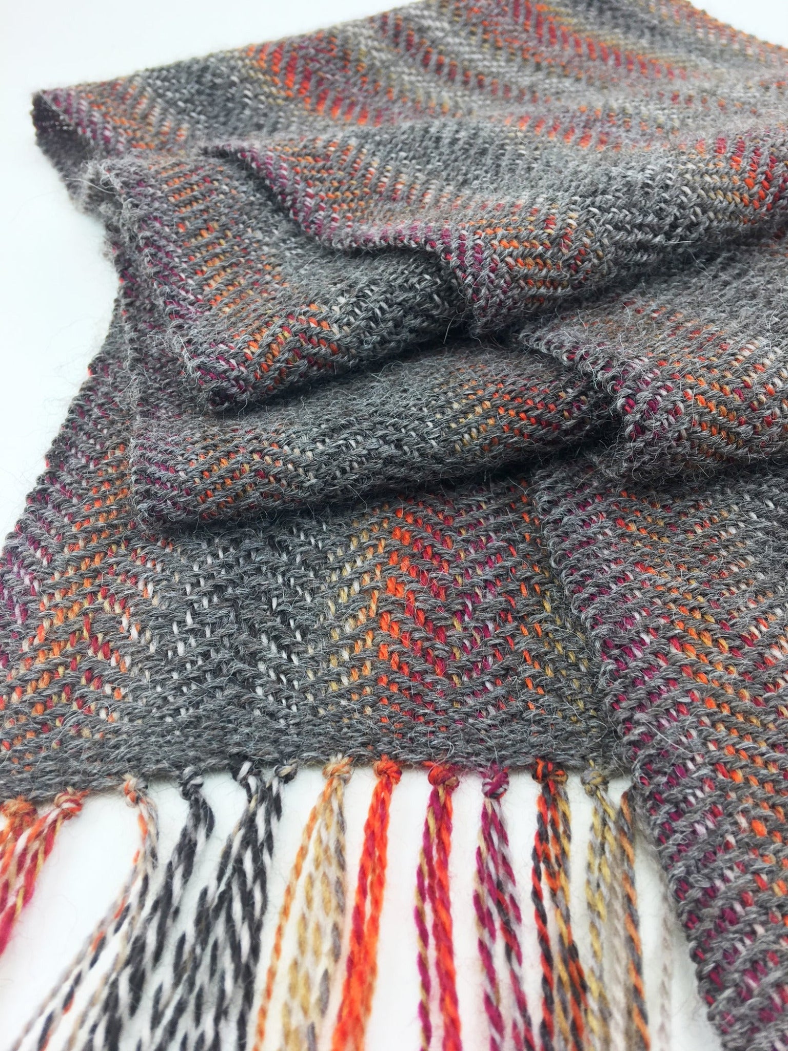 A close-up of a folded Halcyon Yarn Oktoberfest - Woven Scarf, download, showcasing its handwoven gray fabric with colorful threads woven throughout. The scarf features fringes with multicolored strands hanging from the ends in hues of orange, red, purple, and white. The texture appears soft and warm.