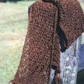 A long Chainette Scarf from Halcyon Yarn drapes elegantly over a rustic wooden rail outside. The brown, tasseled scarf features an open, lacy pattern with tassels at the ends. In the background, a grassy field is slightly out of focus.