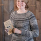 A young person with short blond hair is smiling and holding a book titled "Home Dyeing with Natural Dyes." They are wearing the Mannequin Pullover by Halcyon Yarn, a gray worsted weight sweater featuring a raglan yoke, and standing in front of a wooden plank wall.