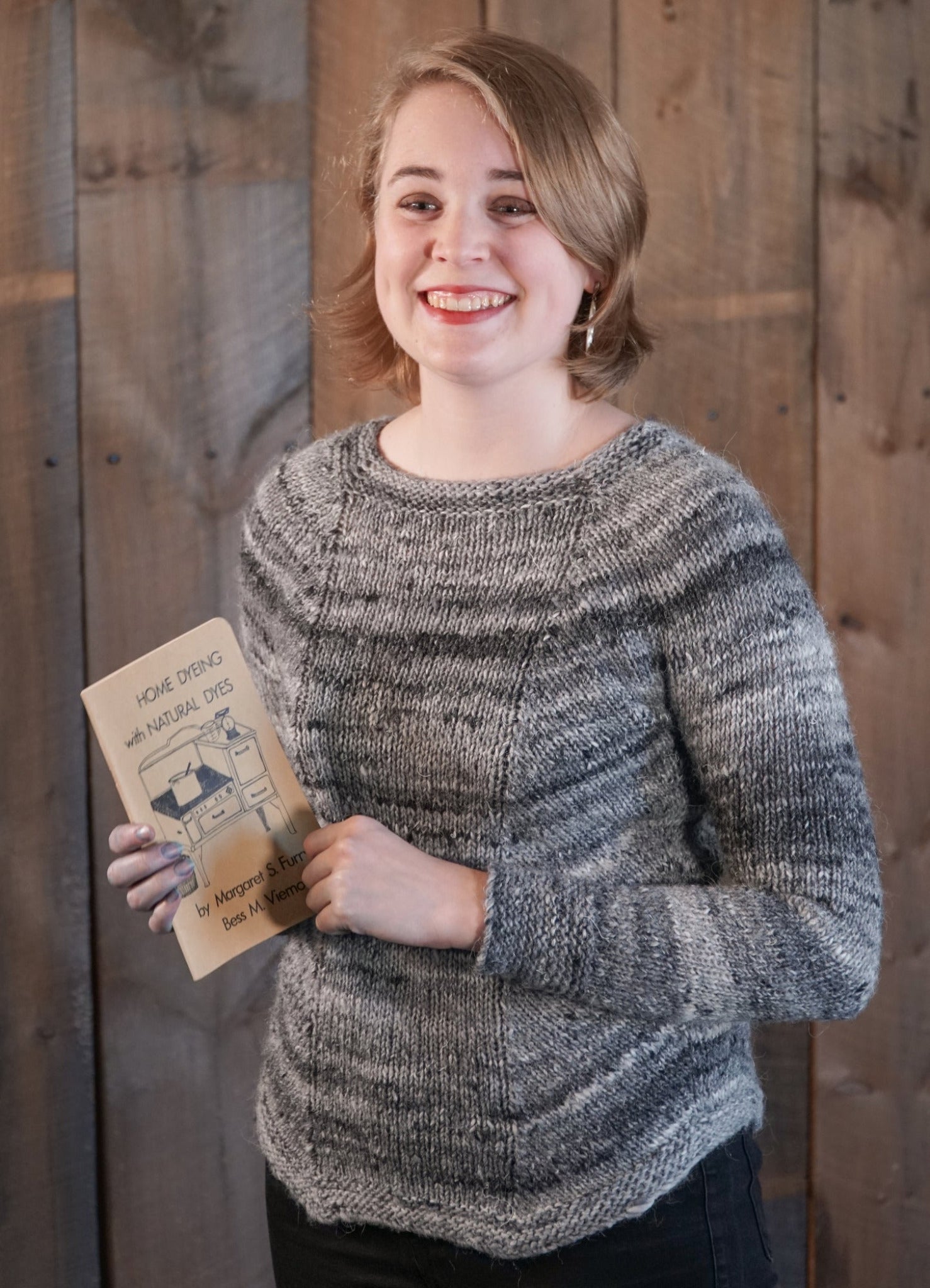 A young person with short blond hair is smiling and holding a book titled "Home Dyeing with Natural Dyes." They are wearing the Mannequin Pullover by Halcyon Yarn, a gray worsted weight sweater featuring a raglan yoke, and standing in front of a wooden plank wall.