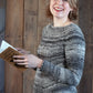 A young woman with short blonde hair smiles brightly while holding an open book. She is wearing the Halcyon Yarn Mannequin Pullover, a gray worsted weight sweater with a raglan yoke, and stands in front of a rustic wooden background.