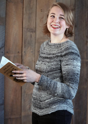 A young woman with short blonde hair smiles brightly while holding an open book. She is wearing the Halcyon Yarn Mannequin Pullover, a gray worsted weight sweater with a raglan yoke, and stands in front of a rustic wooden background.