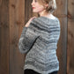 A person with short, light brown hair is turned slightly to the side, looking over their shoulder. They are wearing the Halcyon Yarn Mannequin Pullover, a gray knitted sweater with a raglan yoke, paired with dark pants. They are standing in front of a wooden wall.