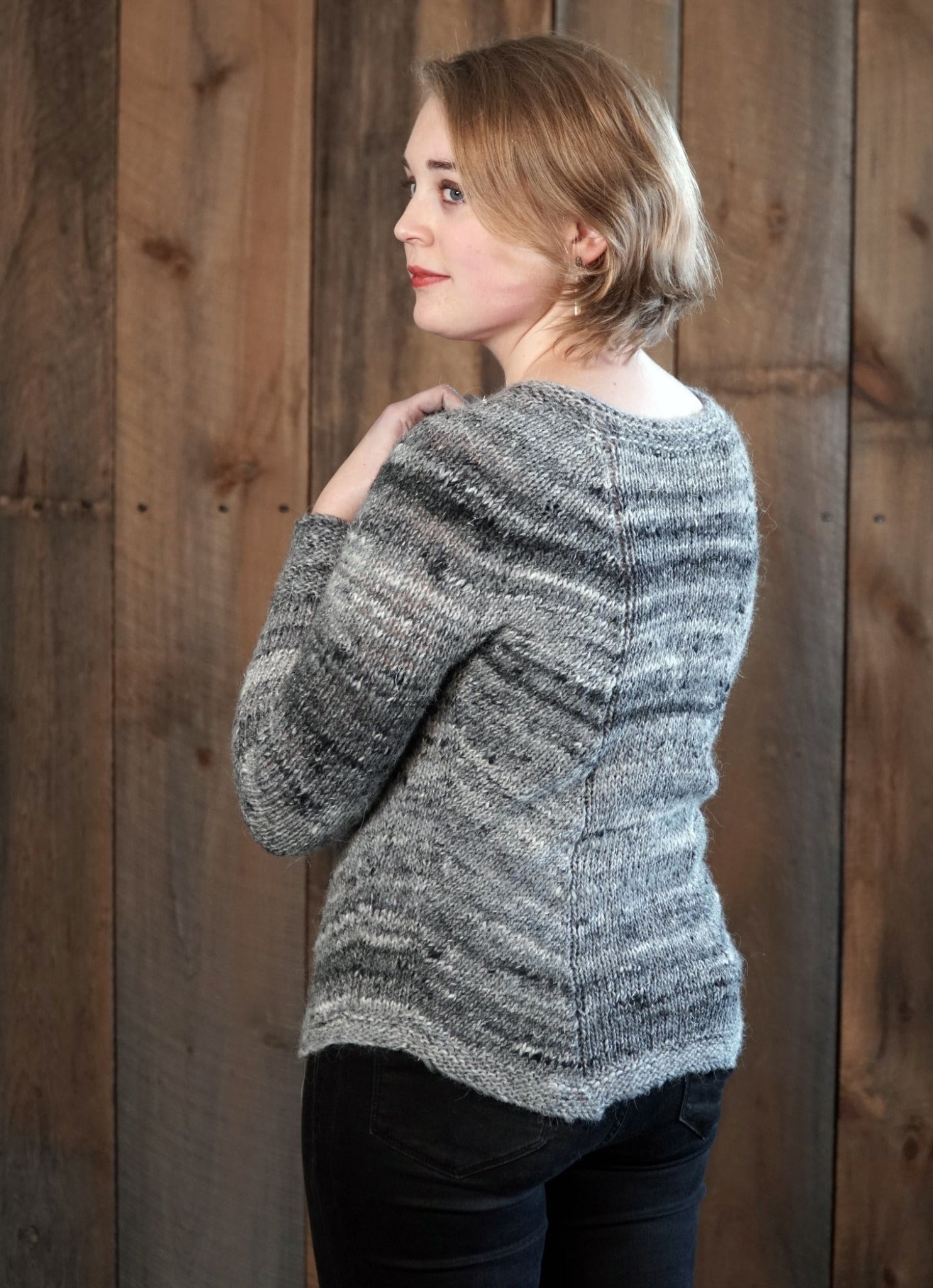 A person with short, light brown hair is standing sideways, looking back over their shoulder. They are wearing the Mannequin Pullover by Halcyon Yarn, a gray worsted weight sweater with a textured pattern, paired with black pants. The background features wooden planks.