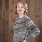 A person with short, light brown hair is smiling and posing with one hand on their hip. They are wearing the Mannequin Pullover, a gray and black worsted weight sweater by Halcyon Yarn. The background is composed of wooden planks.