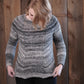 A person with light hair is standing in front of a wooden wall, looking down at the Halcyon Yarn Mannequin Pullover they are wearing. The worsted weight sweater features a gradient pattern from light to dark grey and intricate bias shaping. The person is pulling at the hem of the pullover with both hands.