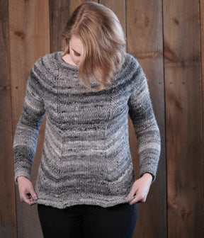 A person with shoulder-length blonde hair is looking down while holding the hem of their grey Mannequin Pullover. The worsted weight sweater, by Halcyon Yarn, features horizontal stripe patterns. The background consists of wooden panels.