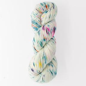 A skein of Happy Feet Sock Yarn from Plymouth Yarn Co. in a cream base color, speckled with vibrant hues of blue, pink, orange, and yellow, is coiled neatly against a white background. Made from superwash Merino wool, this durable knitting yarn's colorful speckles create a visually interesting and playful pattern.