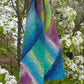 The vibrant Multi-Directional Scarf from Halcyon Yarn, hand-knitted with an array of blue, green, purple, and yellow hues, drapes elegantly over a branch of a blooming tree adorned with white flowers, creating a striking contrast against the natural backdrop. This wonderful project incorporates short rows and is delightful to wear.