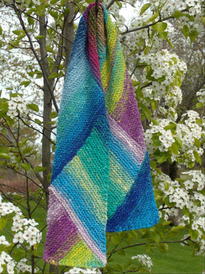 The vibrant Multi-Directional Scarf from Halcyon Yarn, hand-knitted with an array of blue, green, purple, and yellow hues, drapes elegantly over a branch of a blooming tree adorned with white flowers, creating a striking contrast against the natural backdrop. This wonderful project incorporates short rows and is delightful to wear.