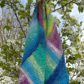 A vibrant Multi-Directional Scarf by Halcyon Yarn, featuring short rows in hues of blue, purple, green, and yellow, drapes elegantly on a blooming tree branch adorned with white flowers against a backdrop of clear skies.